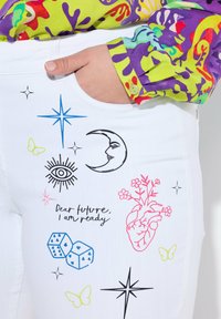 Valkoiset farkut, joissa on värikkäitä graafisia kuvastoja, kuten kuu, silmä, sydän ja kukkia, sekä teksti "Rakas tulevaisuus, olen valmis."