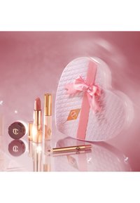 Scatola cosmetica a forma di cuore di un bianco lucido con un fiocco di raso rosa. Include rossetto rosa, lucidalabbra e contorno labbra, con dettagli dorati.