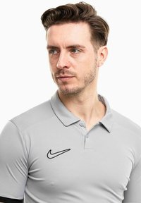Helles graues Poloshirt aus glattem Stoff, mit schwarzem Nike-Logo auf der linken Seite, samt Kragen und Knopfleistendetails.