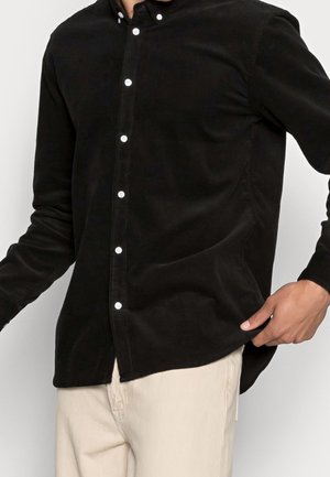 Homme portant une chemise noire à manches longues boutonnée avec des boutons blancs et un pantalon beige clair, tenant l'ourlet de la chemise d'une main.