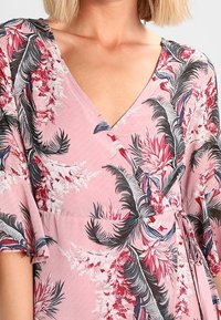 Kwiatowa sukienka w kolorze różowym z krótkimi rękawami. Posiada dekolt w serek, tropikalne wzory liści w kolorach czerwonym, zielonym i czarnym oraz zwężoną talię.