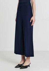 Modella femminile che indossa pantaloni cropped a gamba larga blu navy abbinati a mule nere con punte, caratterizzati da dettagli di occhielli in argento.