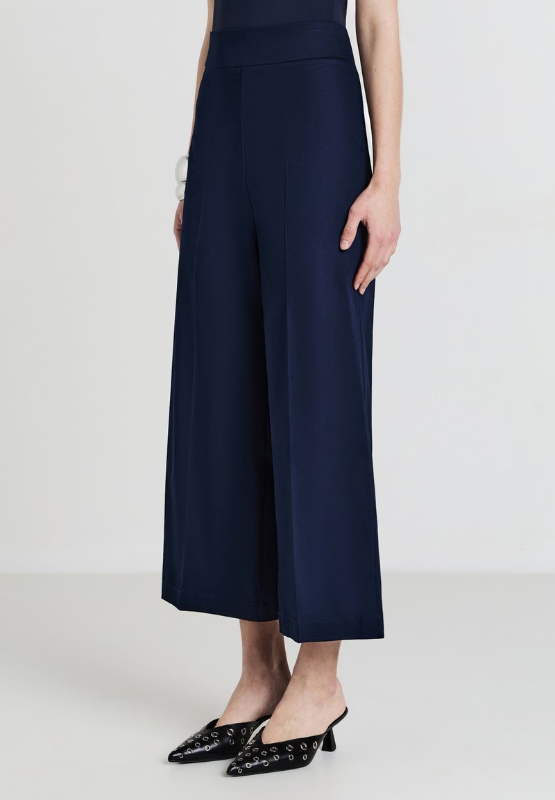Modella femminile che indossa pantaloni cropped a gamba larga blu navy abbinati a mule nere con punte, caratterizzati da dettagli di occhielli in argento.