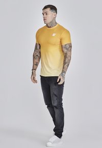 Kortærmet gradient t-shirt, der går fra lys gul til sart gul, parret med sorte ripped jeans og hvide sneakers med teksturerede detaljer.