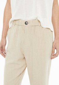 Pantalones beiges a rayas con un cierre de botón en la cintura, que presentan una tela suave y un corte relajado, parcialmente cubiertos por una blusa blanca.