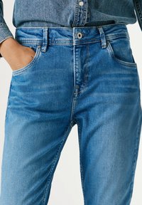 Close-up van een persoon die een blauwe spijkerbroek draagt met de hand in de zak en een bijpassend blauw spijkerhemd dat in de broek is gestopt.