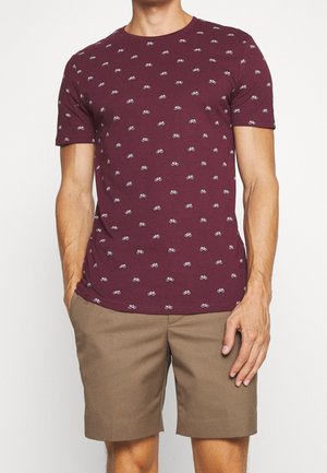 Bordeauxfarbiges Baumwoll-T-Shirt mit einem wiederholten weißen Fahrradmuster, kombiniert mit khakifarbenen Shorts. Kurze Ärmel und Rundhalsausschnitt.