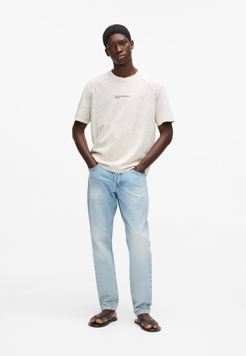 Svetlosivé tričko s textovým logom, v kombinácii s vybledlými modrými džínami a čiernymi žabkami. Krátke rukávy a voľný strih.