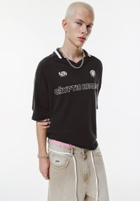 Černý oversized krátký rukávový dres s bílými akcenty a textem "CRYPTIC REALS"; kombinován s volnými kalhotami světlé barvy s bílou šňůrkou na stažení.
