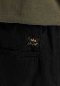 Tissu noir avec une petite étiquette cousue en jaune indiquant "Alpha Industrie Knoxville, Tennessee USA" au-dessus d'une couture de poche.