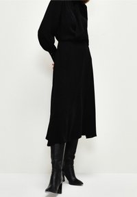 Robe midi noire avec une silhouette fluide, un tissu texturé et un ourlet arrondi. Associée à des bottes noires au-dessus du genou avec un talon en bloc.