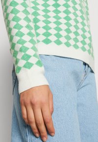 Maglione a motivo romboidale verde e bianco con polsini a costine, abbinato a jeans di denim azzurro chiaro. Tessuto morbido e design aderente.