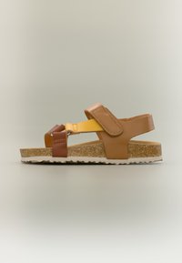 Καφέ, κίτρινο και μπεζ σανδάλι με ρυθμιζόμενα λουριά, πάτο φτιαγμένο από φελλό και καουτσούκ σολάκι, εμφανιζόμενο σε πλαϊνή όψη σε ουδέτερο φόντο.