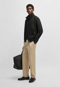 Schwarze Bomberjacke mit Reißverschluss, Stehkragen, zwei seitlichen Taschen, kombiniert mit beigen Hosen und schwarzen Schuhen, hält eine schwarze Tasche.