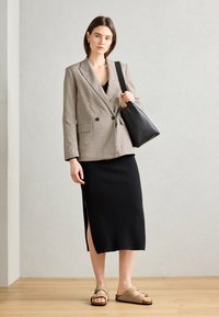 Jupe midi noire à côtes avec une fente sur le côté, associée à un blazer beige à carreaux croisé et des sandales marron. Sac à main noir porté sur l'épaule.