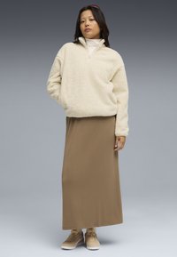 Creme fleece pullover med en halv-zip krave, parret med en lang brun nederdel og beige sneakers, sat mod en grå baggrund.