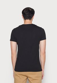 Černé tričko s krátkým rukávem, kulatým výstřihem, hladkým materiálem a rovnými lemy. Nalezeno v kombinaci s béžovými kalhotami, pohled zezadu.