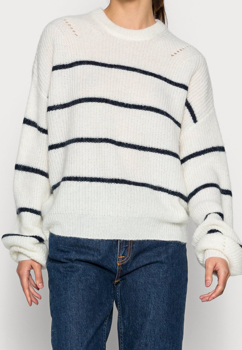 Pull en maille blanche avec rayures horizontales bleu marine, encolure ronde, coupe ample et poignets côtelés. Présente des ouvertures ajourées sur les épaules.
