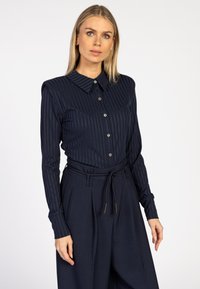 Vrouw met steil blond haar die een marineblauw gestreept overhemd met knopen draagt, gecombineerd met hoge marineblauwe wijde broek met een gevlochten riem.