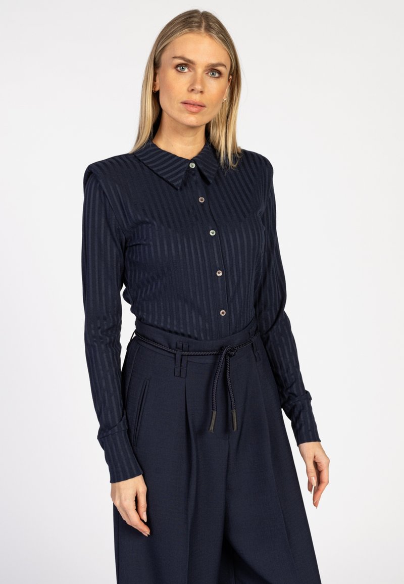 Vrouw met steil blond haar die een marineblauw gestreept overhemd met knopen draagt, gecombineerd met hoge marineblauwe wijde broek met een gevlochten riem.