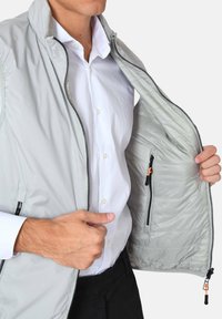 Giacca leggera grigia con zip frontale e tasche, caratterizzata da una superficie liscia e dettagli interni trapuntati.