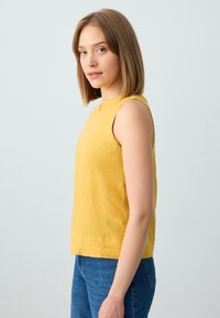 Top giallo senza maniche realizzato in tessuto texturizzato, con scollatura alta e vestibilità ampia, abbinato a jeans blu.