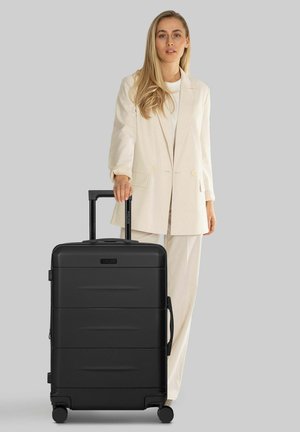 Valigia nera rigida con superficie texturizzata, maniglia retrattile e quattro ruote, posizionata accanto a una persona con un outfit color crema.