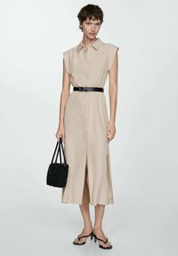 Abito camicia beige con maniche corte, una cintura e bottoni sul davanti. Design di lunghezza media con una fessura laterale. Borsa intrecciata nera e sandali.