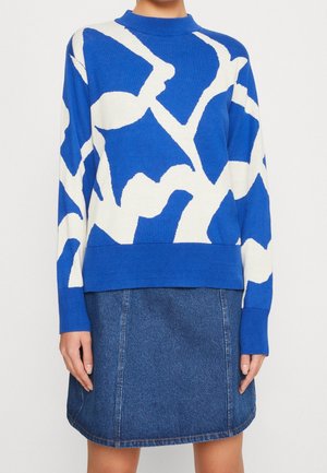 Pull bleu avec des motifs abstraits blancs, à manches longues et col haut, associé à une jupe en denim. Le pull présente des poignets et un ourlet côtelés.