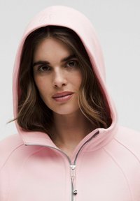 Helle rosa Kapuzenjacke mit Reißverschluss, gerippter Textur und dezentem Logo auf der Kapuze. Verfügt über ein tailliertes Design und weichen Stoff.