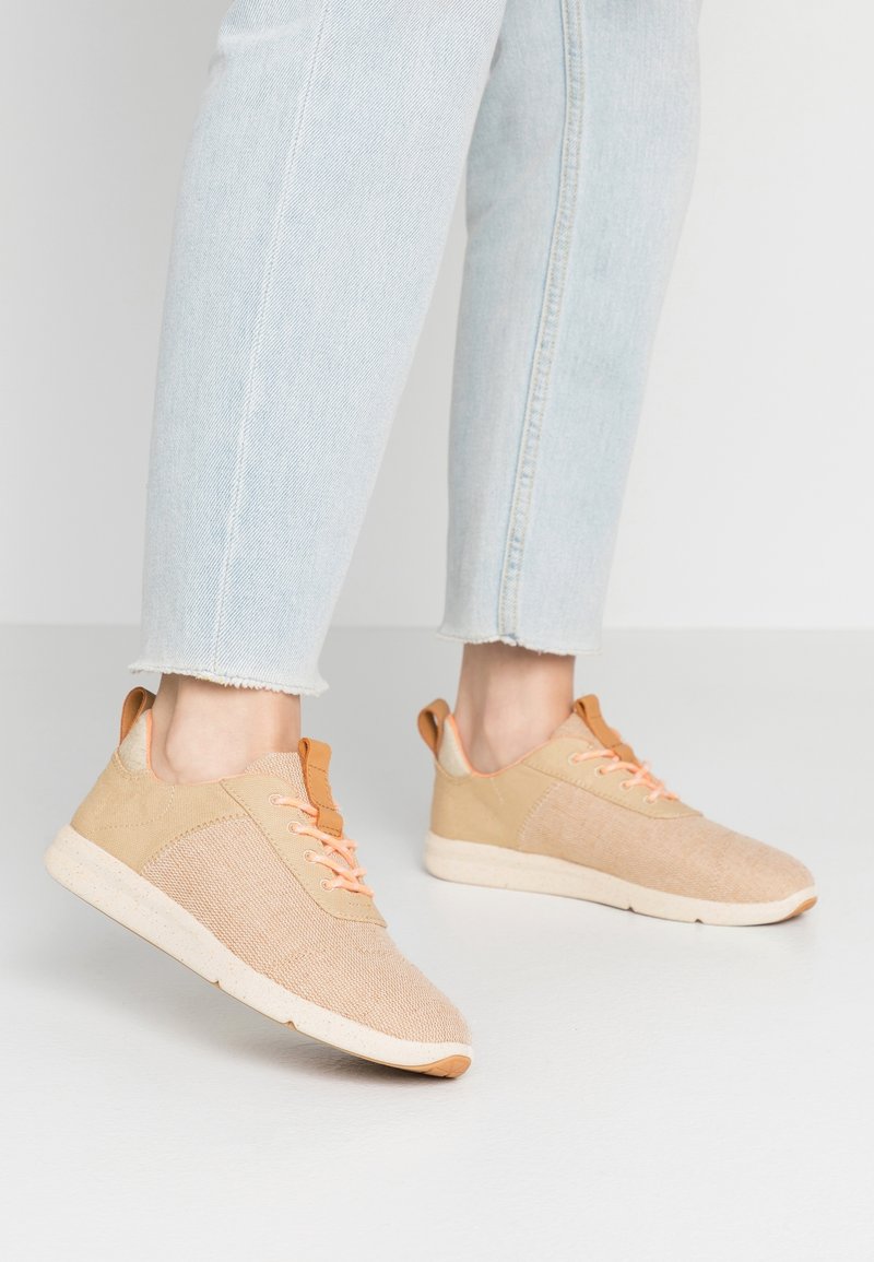 Beige Sneaker mit einem strukturierten Stoff-Obermaterial, orangefarbenen Schnürsenkeln und einer hellbraunen Sohlen. Ausgestattet mit einem gepolsterten Kragen und einem minimalistischen Design.