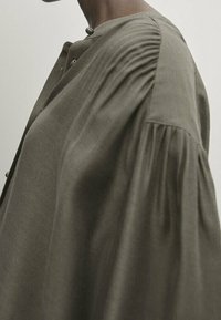 Olive green blouse with a round neckline, featuring gathered detail on the shoulder and flowing, wide sleeves in a soft fabric.