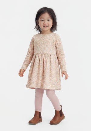 Young girl smiling, wearing a floral long-sleeve dress, pink leggings, and brown ankle boots standing on a white background.
