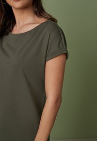 Femme portant une chemise à manches courtes olive avec des revers, se tenant devant un fond vert assorti, montrant le haut du torse et une partie du visage.