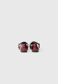 Un paio di scarpe piatte lucide di un rosso scuro con cinturini neri a punta, mostrate dal retro su uno sfondo bianco semplice.