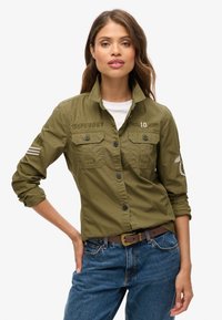 Groene katoenen shirt met lange mouwen, knoopsluiting, borstzakken en geborduurde details. Gepaard met blauwe jeans en een bruine riem.