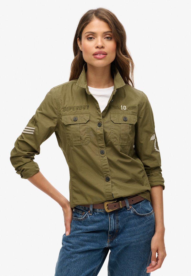 Groene katoenen shirt met lange mouwen, knoopsluiting, borstzakken en geborduurde details. Gepaard met blauwe jeans en een bruine riem.