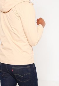 Light beige hooded jacket with a smooth texture, featuring seams and a front zipper, paired with dark denim jeans showing back pocket detail.