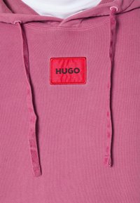 Różowa bluza z kapturem, wykonana z miękkiego materiału, z czerwoną łatą z napisem "HUGO" w czarnym kolorze oraz sznurkami do regulacji kaptura.