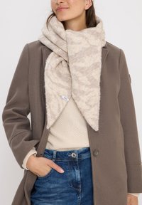 Écharpe beige et crème à motifs en tissu doux et texturé ; de forme triangulaire, elle se drape autour du cou, superposée à un pull léger.