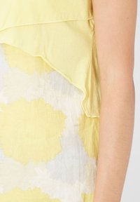 Top amarillo en capas con un dobladillo con volantes, que presenta grandes patrones florales en amarillo claro y blanco. Tejido suave y ligero con una textura suave.