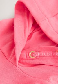 Nahaufnahme eines pinkfarbenen Hoodies mit Kapuze, pinker Kordel mit goldenen Ösen und dezentem Marken-Schriftzug auf der Kordel.
