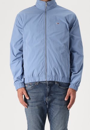 Mann trägt hellblaue Zip-up-Jacke mit elastischen Bündchen und dunkelblaue Jeans, steht mit entspannten Händen an den Seiten.