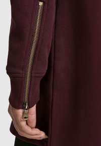 Bordo majica z bogato teksturo, ki ima zlati zadrgo na strani in rebraste manšete. Tkanina je videti mehka in zračna.