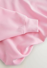 Svetloružová látka mikiny s hladkou textúrou a stiahnutou manžetou. Mäkký materiál sa jemne zvažuje, čím vyzdvihuje jednoduchý a čistý dizajn.