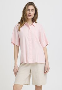 Femme portant une chemise à manches courtes rose clair boutonnée et un short beige à hauteur des genoux, se tenant devant un fond blanc uni.