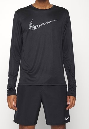 Osoba nosząca czarną koszulkę z długim rękawem z wzorzystym białym logo Nike oraz czarne szorty Nike z białym logo na lewej nodze.
