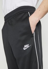 Osoba v černých Nike teplákových kalhotách s bílými postranními pruhy a logem, ruka v kapse, doplněná bílým tričkem.