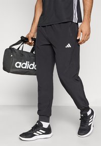 Sorte atletikbukser med lynlåslommer og elastiske manchetter, kombineret med sorte og hvide sneakers og en Adidas duffel-taske.