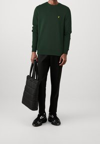 Sudadera verde oscuro con logo amarillo, pantalones negros y bolso tote acolchado negro. El modelo lleva zapatillas deportivas negras. Diseño sencillo y moderno.